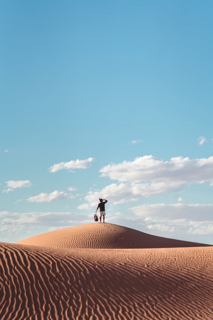 A lone figure stands atop a sand dune under a vast blue sky in the Egyptian desert.